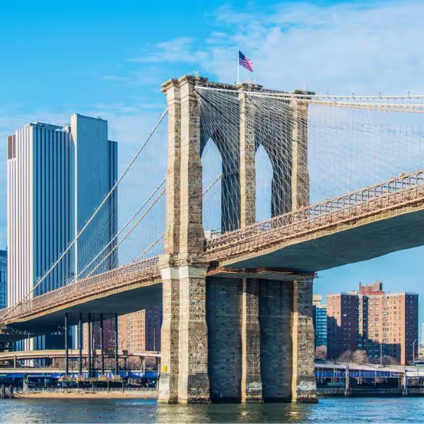 image of Brooklyn Bridge with American flag on top and blue skies and buildings in background